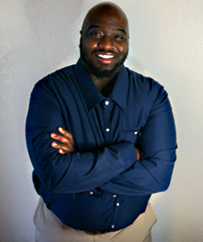 This photo contains a smiling, Black man with a bald head, beard, and brown eyes. He is wearing a royal blue long sleeve dress shirt with white buttons, light khaki pants, and has his arms crossed. He is standing against a white background.