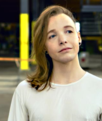 The photo contains a non binary, trans person with a dreamy expression who has blue/grey eyes, shoulder length blonde hair that’s swept to one side and a white t-shirt on.